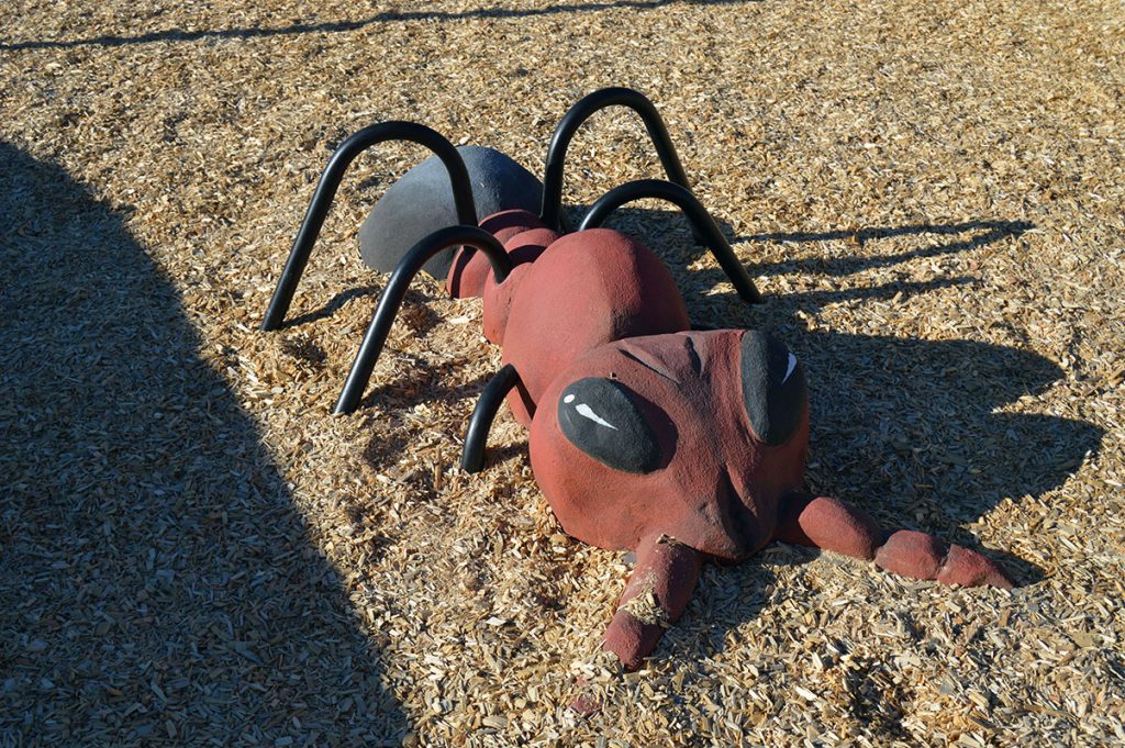 Large red and black ant sculpture on a wood chip playground surface.