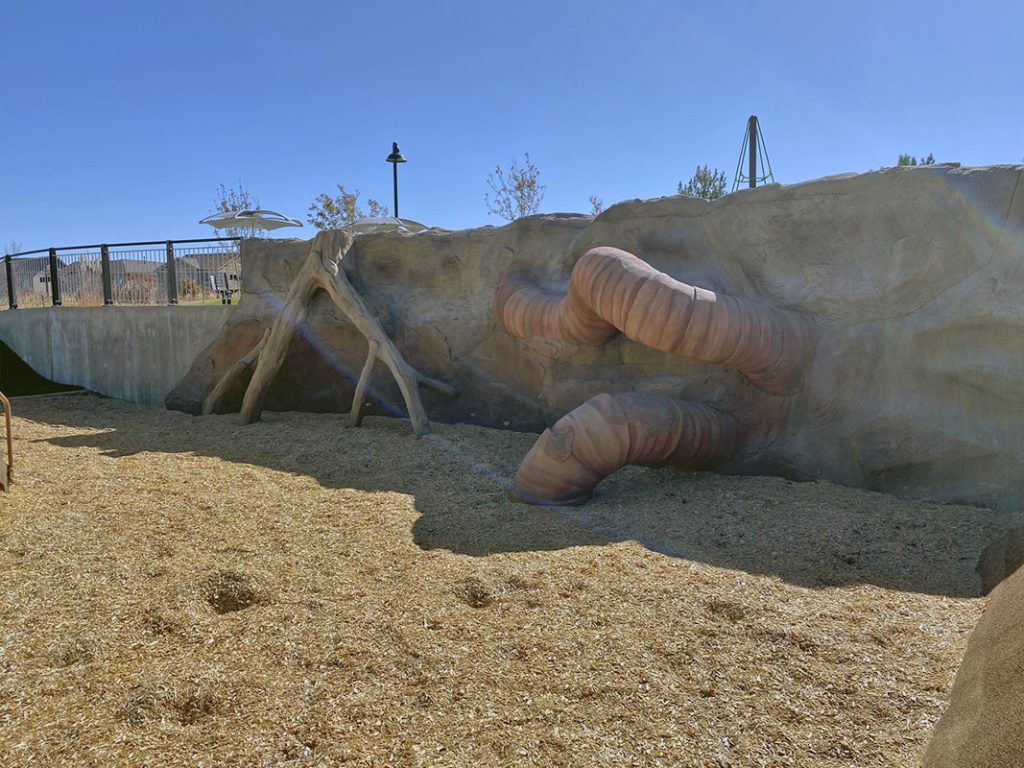 A playground with a rock-like wall featuring cylindrical, earthworm-like protrusions and wood chip ground cover.