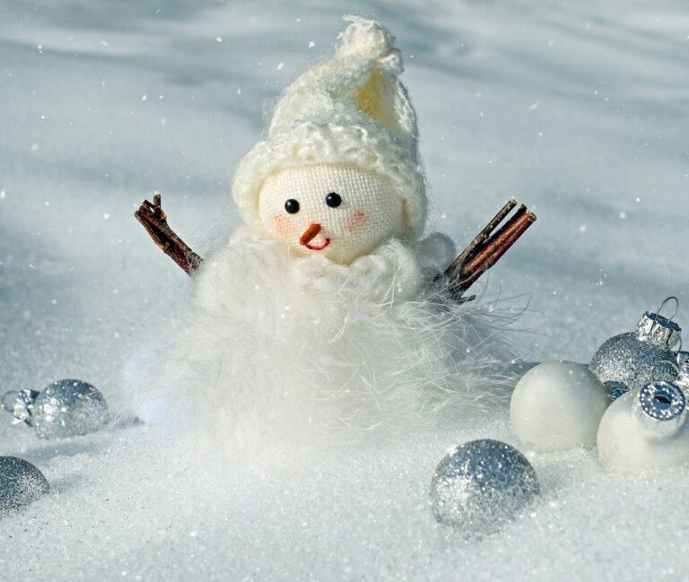 Decorative snowman with twigs for arms in snow, surrounded by silver and white Christmas ornaments.